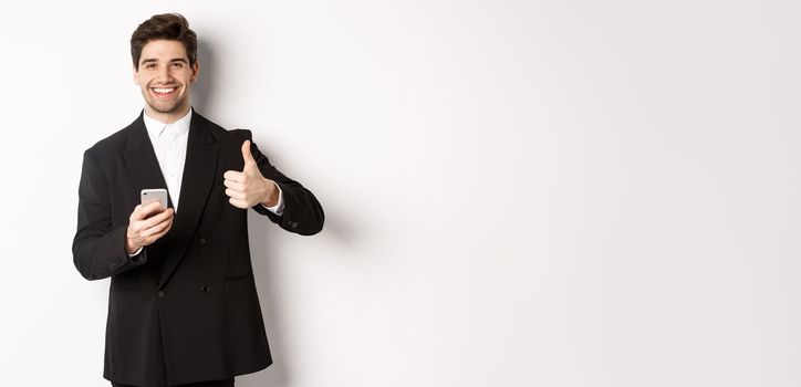 Handsome guy in trendy suit making compliment, showing thumbs-up while using mobile phone, recommending an app or online shop, standing over white background.