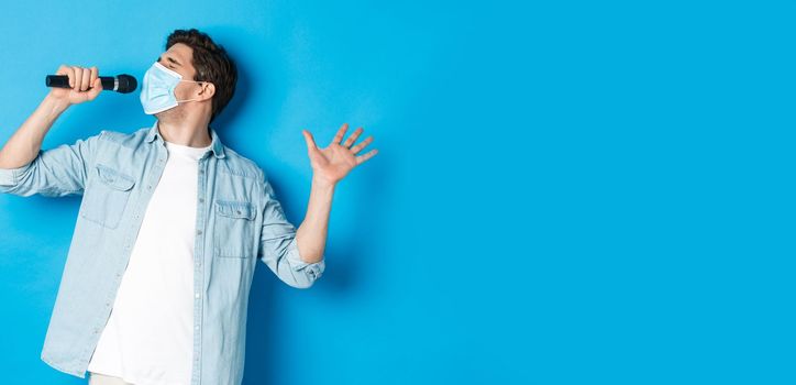 Handsome guy in medical mask singing in microphone, standing over blue background.