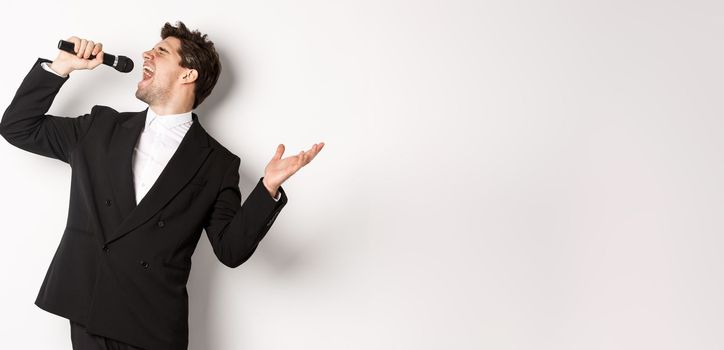 Portrait of handsome man singing a song with passion, standing in black suit, holding microphone and performning, posing over white background.