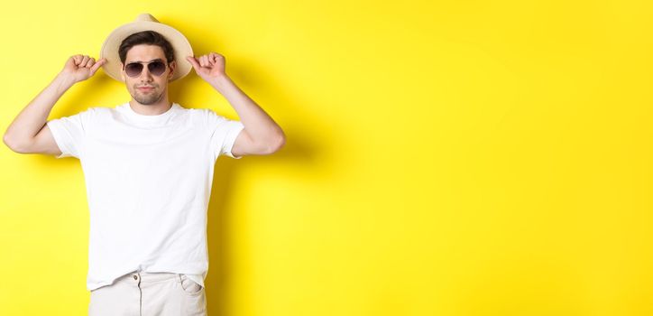 Confident young male tourist ready for vacation, wearing straw hat and sunglasses, standing against yellow background.