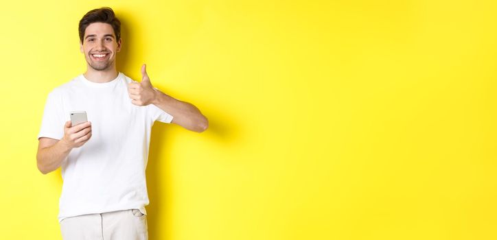 Happy satisfied man holding smartphone, showing thumb up in approval, recommend something online, standing over yellow background.