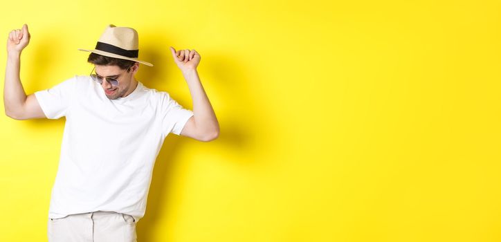 Tourism, travelling and holidays concept. Man tourist enjoying vacation, dancing in straw hat and sunglasses, posing against yellow background.