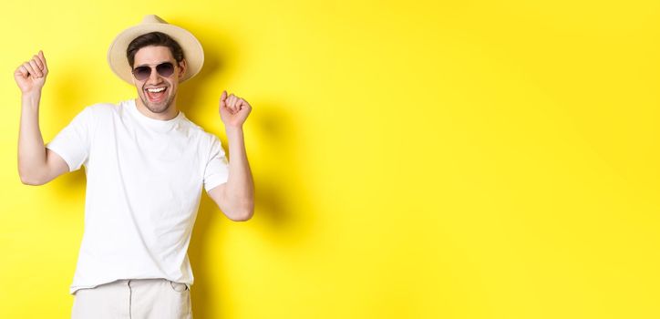 Tourism, travelling and holidays concept. Happy caucasian guy dancing and having fun on vacation, wearing sunglasses with straw hat, standing against yellow background.