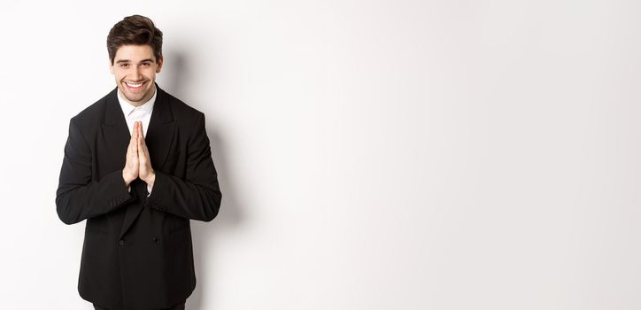 Portrait of handsome man in black suit, being grateful, saying thank you and bowing politely, smiling as holding hands together, expressing gratitude, standing over white background.