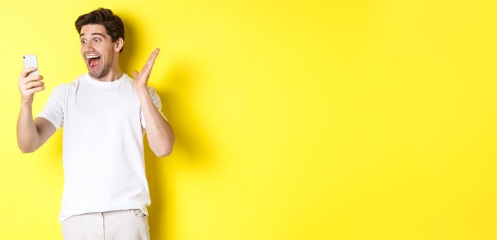 Surprised and happy man looking at mobile phone screen, reading fantastic news, standing over yellow background.