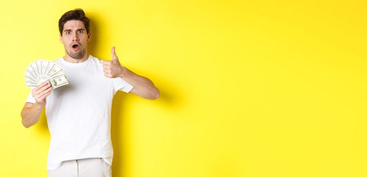 Impressed man showing thumb up, holding money credit, standing over yellow background.