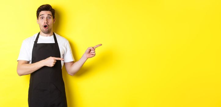 Amazed waiter in black apron showing promo offer, pointing fingers right and looking surprised, standing in uniform against yellow background.