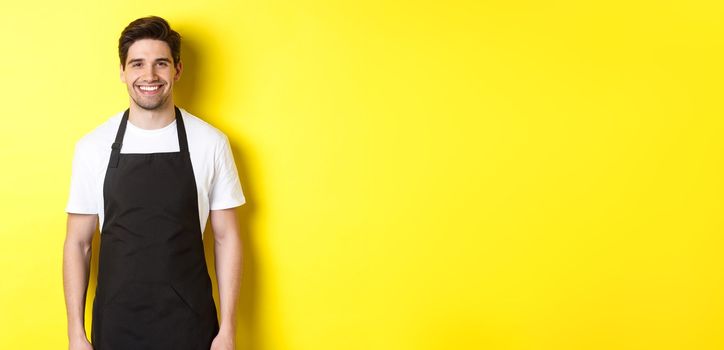 Young man waiter in black apron, smiling, working in store or coffee shop, standing against yellow background.