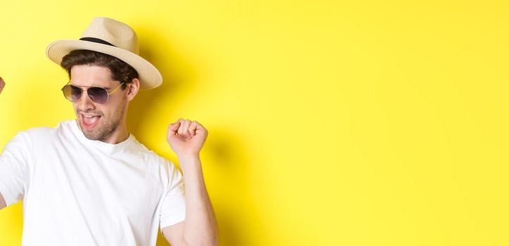 Concept of tourism and vacation. Close-up of man enjoying holidays on trip, dancing and pointing fingers sideways, wearing sunglasses with straw hat, yellow background.