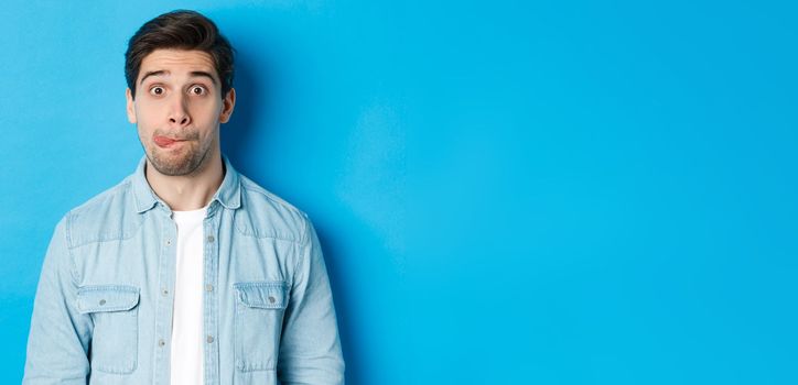 Close-up of young man making funny expressions, showing tongue and looking at camera, standing over blue background.