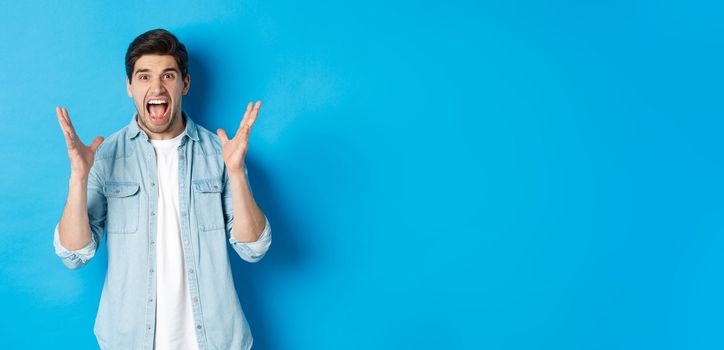 Frustrated young man screaming and looking tensed, shaking hands mad, standing against blue background.