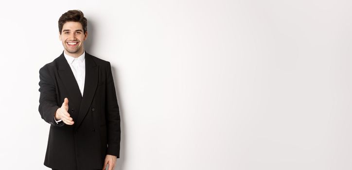 Portrait of handsome businessman in black suit, extending hand for handshake, greet business partners and smiling, welcome to company, standing over white background.