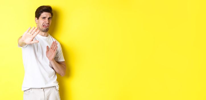 Disgusted man refusing, grimacing from dislike and aversion, begging to stop, standing in white t-shirt against yellow background.