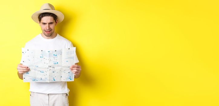 Travelling, vacation and tourism concept. Man looking confused at map during trip, cant understand, standing over yellow background.