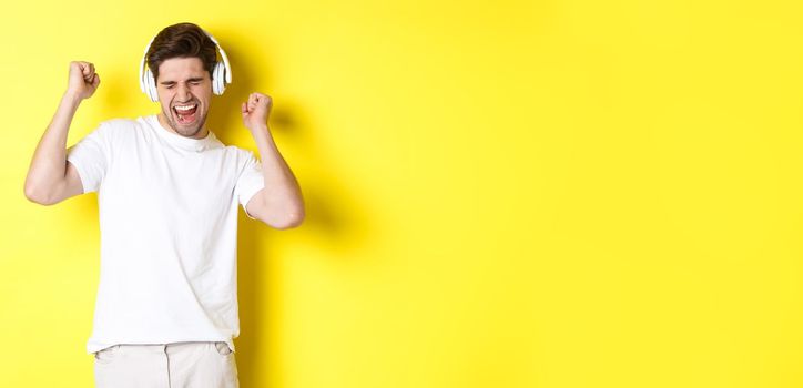 Happy man dancing and listening music in white headphones, standing over yellow background. Copy space