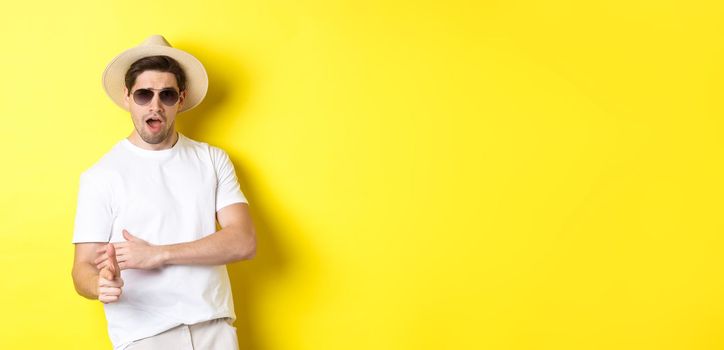 Handsome guy making finger gun shot and looking sassy, dressed for summer vacation in straw hat and sunglasses, standing against yellow background.