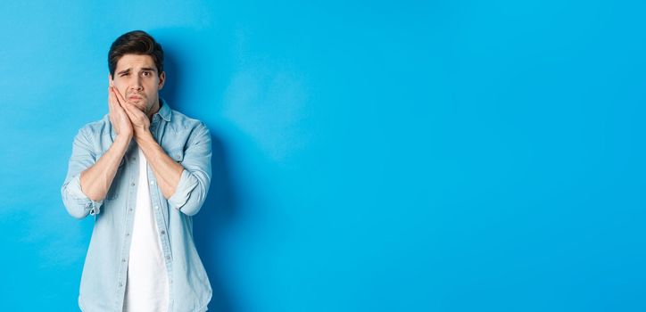 Portrait of man with toothache grimacing from pain and touching cheek, standing against blue background.