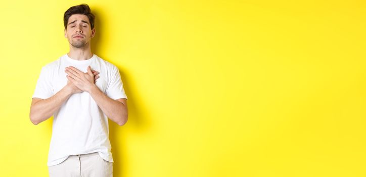 Romantic and nostalgic man holding hands on heart, close eyes and remember something, standing over yellow background.