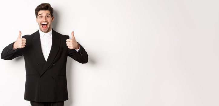 Concept of new year party, celebration and lifestyle. Portrait of amazed and pleased handsome man in black suit, showing thumbs-up, like product, approve something good, white background.