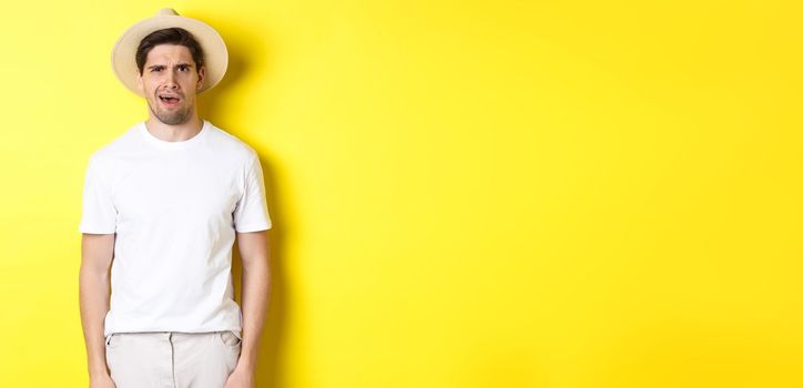 Concept of tourism and summer. Confused guy traveller in straw hat, looking puzzled, cant understand something, standing over yellow background.