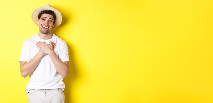 Concept of tourism and summer. Grateful guy in straw hat holding hands on heart, saying thank you and smiling with gratitude, standing against yellow background.