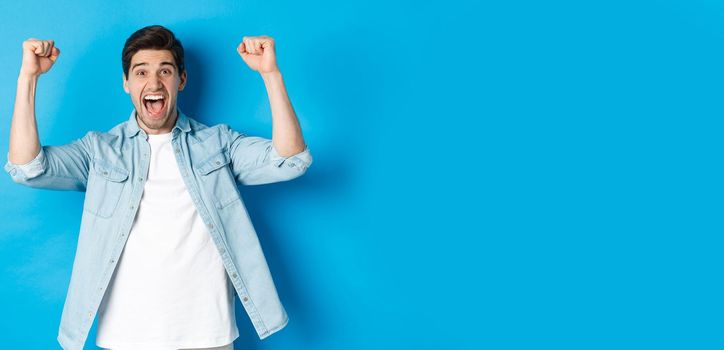 Happy attractive man triumphing, screaming yes and raising hands up to celebrate win, achieve goal, standing against blue background.