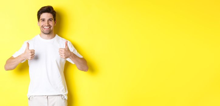 Portrait of happy man showing thumbs-up in approval, like something or agree, standing over yellow background.