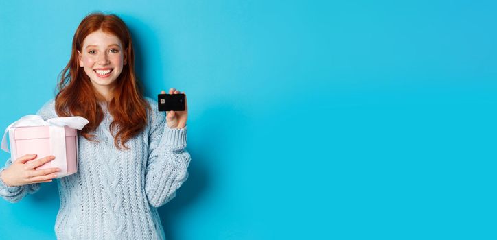 Happy redhead girl buying gifts with credit card, holding box with present and smiling, standing over blue background.