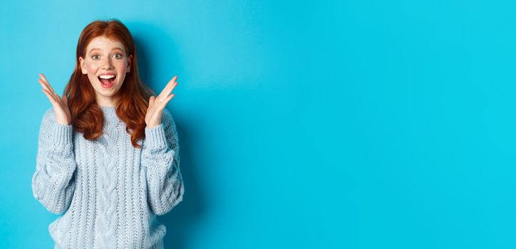 Surprised and happy redhead girl clap hands and staring at camera, smiling amazed, standing against blue background.