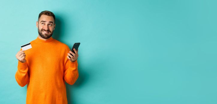 Online shopping. Thoughtful man holding credit card and mobile phone, thinking of purchase, standing over light blue background.