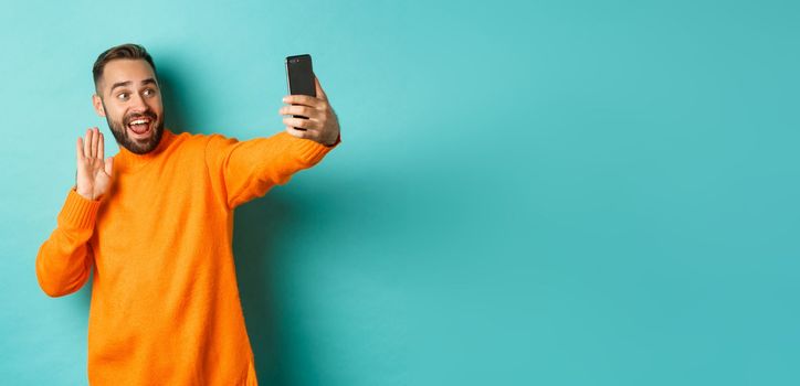 Happy young man video calling, talking online with mobile phone, saying hello to smartphone camera and waving hand friendly, standing over light blue background.