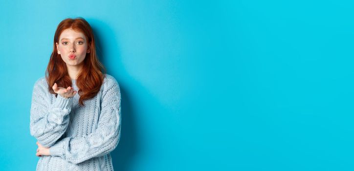 Lovely teen girl in sweater blowing air kiss, pucker lips and staring at camera, standing against blue background.