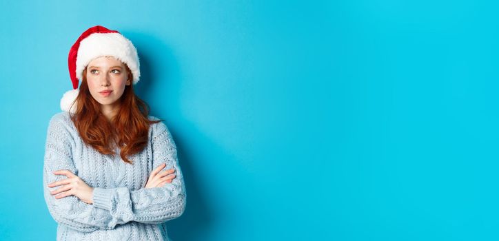 Winter holidays and Christmas Eve concept. Thoughtful redhead woman in Santa hat and sweater, looking left and pondering, making xmas plans, standing over blue background.