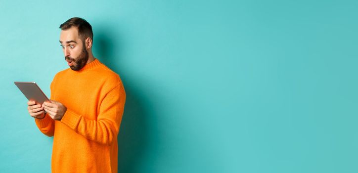 Image of male model in orange sweater staring at digital tablet screen, looking surprised, standing over light blue background.