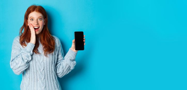 Amazed redhead girl looking at camera, showing smartphone screen, demonstrating mobile application, standing over blue background.