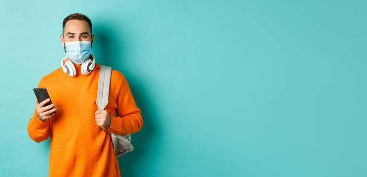 Young man in face mask using mobile phone, holding backpack, standing against light blue background. Copy space