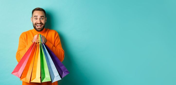 Excited adult man holding shopping bags and smiling, going to mall, standing over turquoise background.