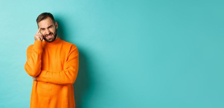 Man with beard, grimacing and cringe from something embarrassing, looking with awkward face, standing over light blue background.