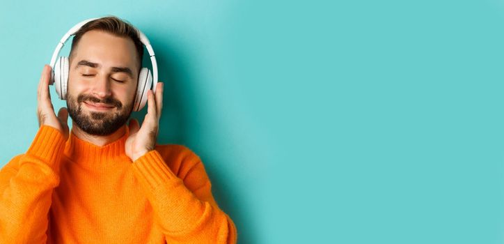 Close-up of handsome modern man listening music in headphones, standing in orange sweater over turquoise background.