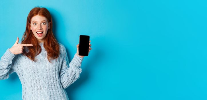 Impressed redhead girl pointing at phone screen, showing smartphone app or online offer and smiling excited, standing in sweater against blue background.