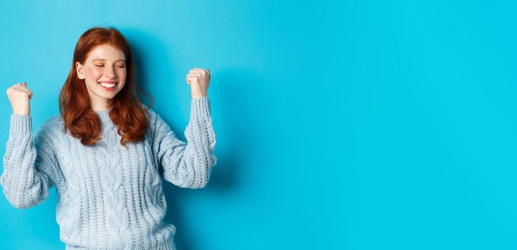 Satisfied redhead girl achieve goal and celebrating, making fist pump gesture and smiling with rejoice, triumphing of win, standing against blue background.