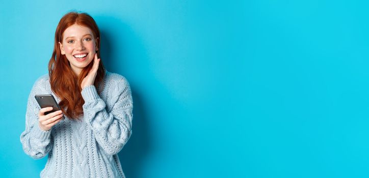 Beautiful redhead girl in sweater, smiling at camera, using mobile phone app, standing with smartphone against blue background.