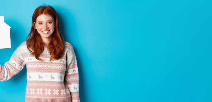 Real estate concept. Young smiling woman with red hair showing paper house model, standing over blue background.