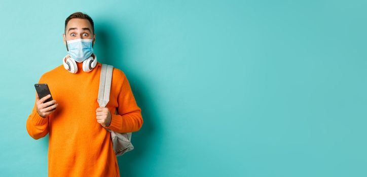 Young man in face mask using mobile phone, holding backpack, staring impressed at camera, standing against light blue background.