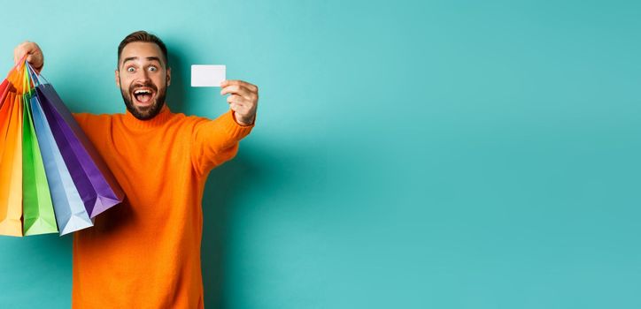 Happy aduly man showing credit card and shopping bags, standing against turquoise background.