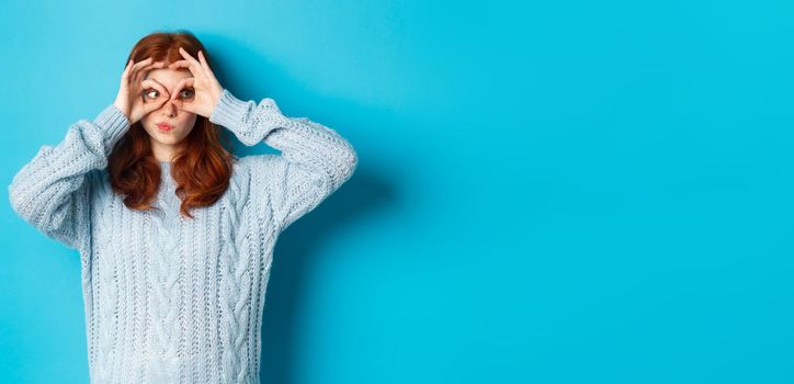 Funny redhead female model in sweater, staring at camera through fingers glasses, express interest and amazement, standing over blue background.