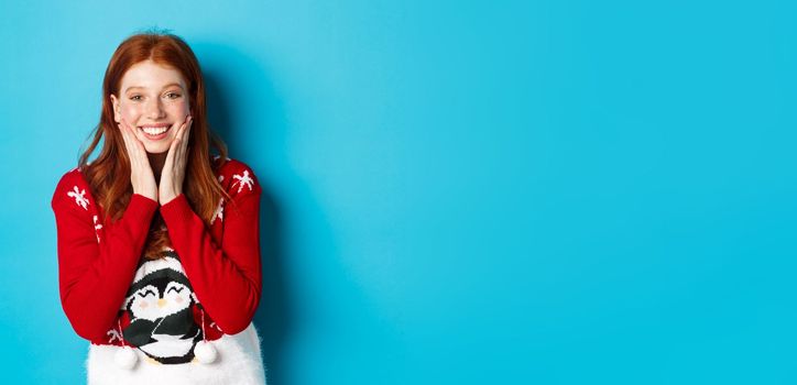 Winter holidays and Christmas Eve concept. Adorable redhead girl blushing and touching cheeks, staring grateful and happy at camera, standing in xmas sweater against blue background.