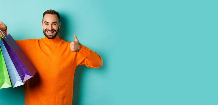 Happy handsome man holding shopping bags and smiling, showing thumbs-up and recommending store, standing over turquoise background.