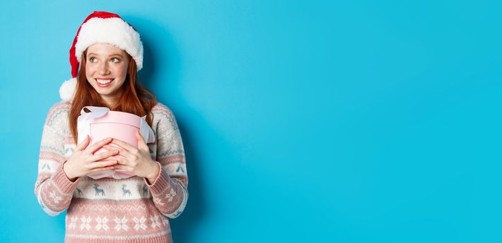 Winter and celebration concept. Dreamy redhead girl in santa hat hugging her christmas gift and looking left, smiling happy, standing over blue background.