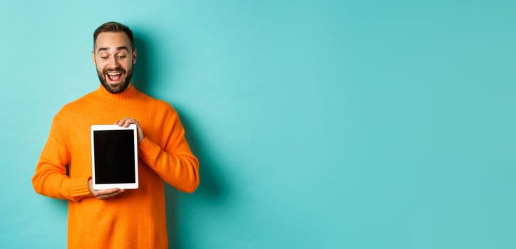 Technology. Cheerful adult man looking and showing digital tablet screen, standing over light blue background.